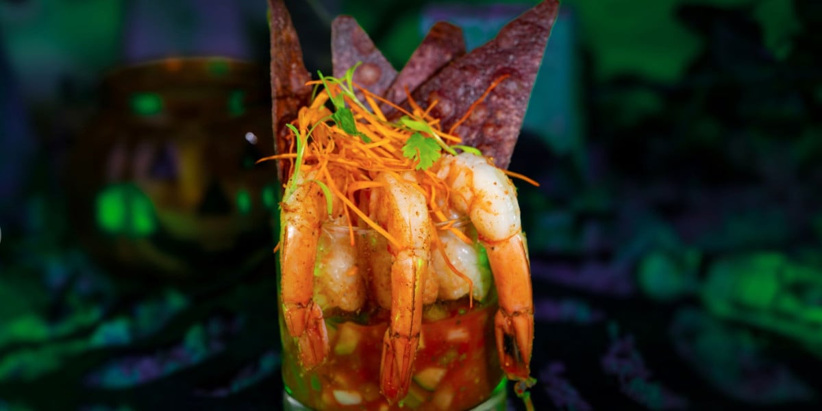 A glass filled with shrimp cocktail, garnished with shredded carrots and cilantro, is served with triangular tortilla chips in the back—a dish that brings a touch of Disney magic to a vibrant, green, out-of-focus background.