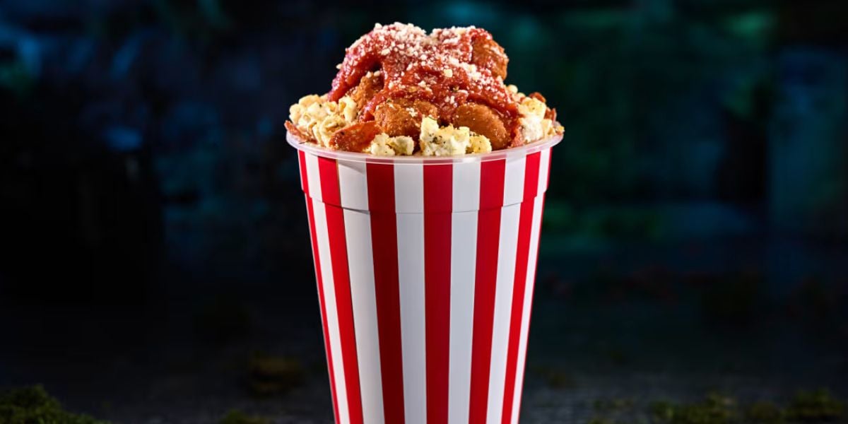 A red-and-white striped tub filled with popcorn, topped with fried chicken pieces, marinara sauce, and grated cheese—perfect for horror nights—set against a dark, blurred background.