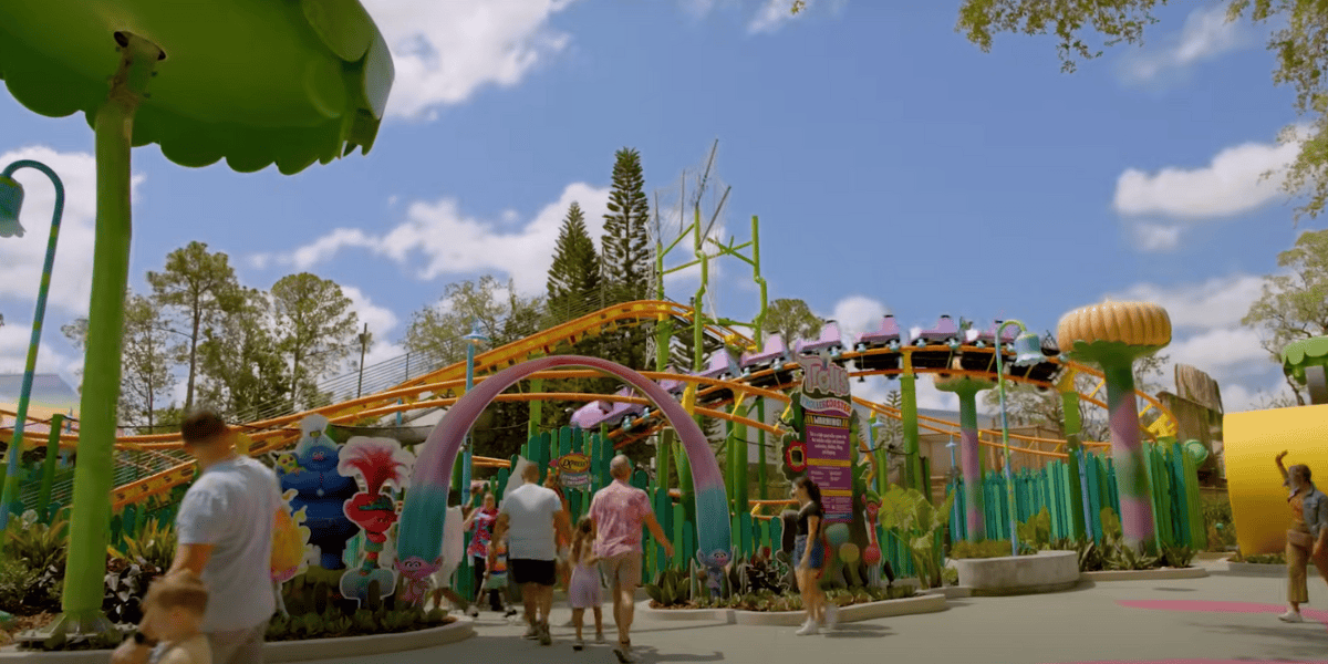 Families walk through a colorful, whimsical amusement park with large playful decorations and a roller coaster above, enjoying a universal sense of wonder under a bright blue sky with scattered clouds.
