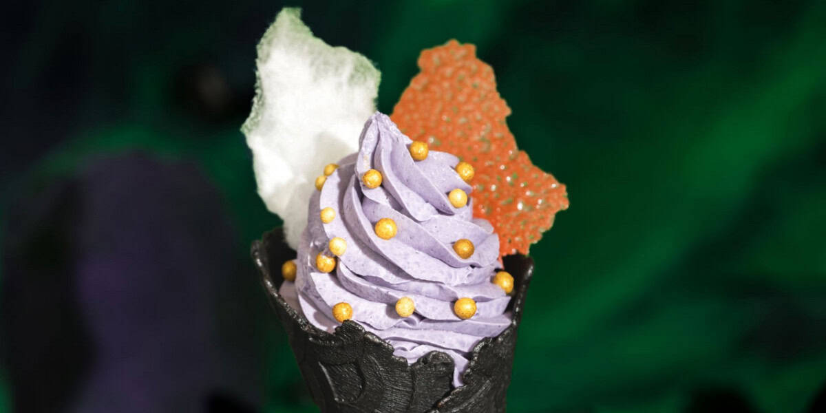 A black waffle cone holds swirled purple soft serve topped with gold candy pearls, a piece of white cotton candy, and an orange sugar decoration—a Disney-inspired treat—set against a dark green background.