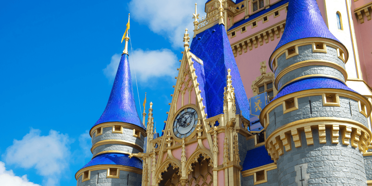 Close-up of a colorful fairytale castle in Magic Kingdom, with blue pointed roofs, gold accents, gray stone walls, and a clock above the entrance, set against a bright blue sky with scattered white clouds at Walt Disney World.