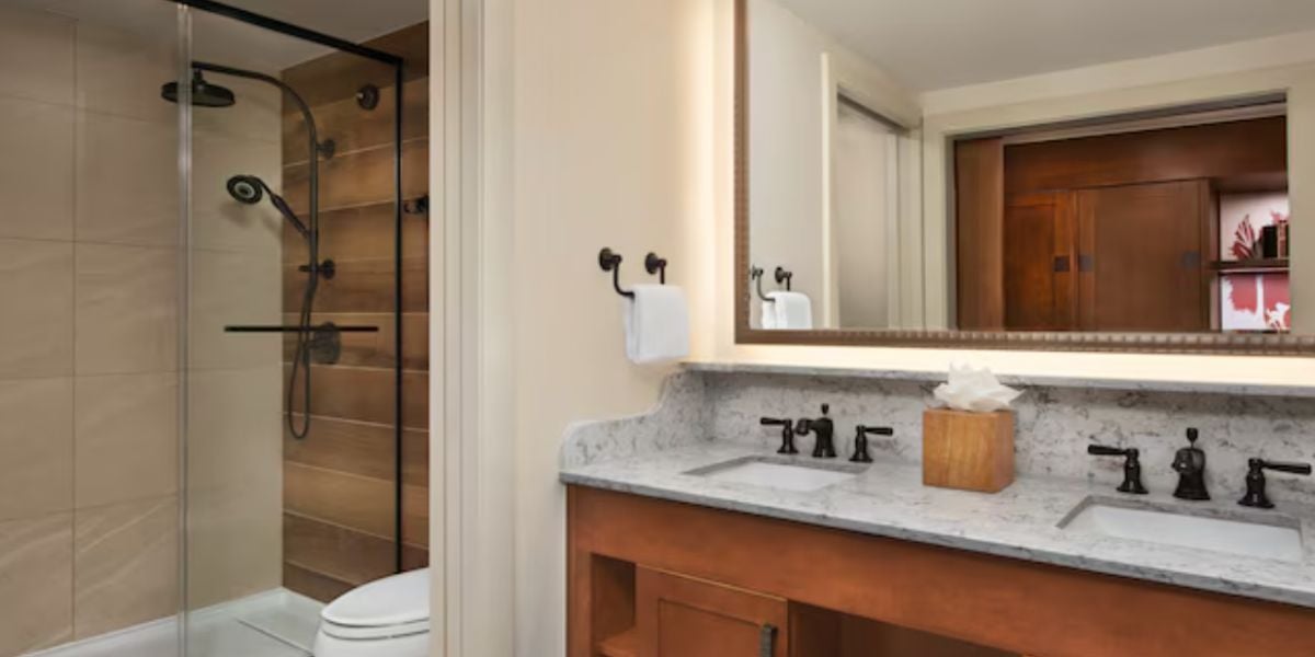 Modern bathroom with a double-sink vanity, marble countertop, large mirror, and a wooden tissue box—bringing the warmth of a wilderness lodge indoors. To the left is a glass-enclosed shower with beige tiles and a black fixture, next to a toilet.