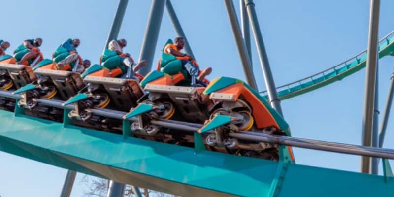 People riding a green and orange Six Flags roller coaster climb up the track, surrounded by steel beams and a blue sky. The excited riders are clearly enjoying this thrilling amusement park experience.