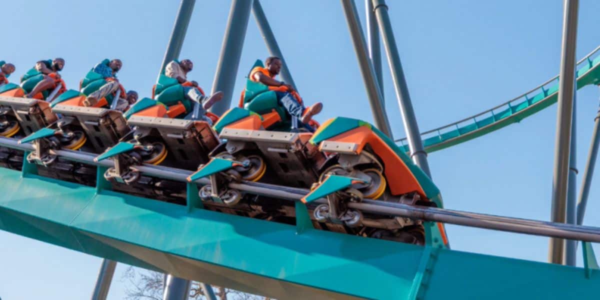 People riding a green and orange Six Flags roller coaster climb up the track, surrounded by steel beams and a blue sky. The excited riders are clearly enjoying this thrilling amusement park experience.
