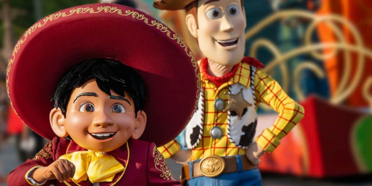 A person in a large Miguel costume from "Coco" stands beside someone dressed as Woody from "Toy Story" at a colorful parade, creating a lively and festive atmosphere.