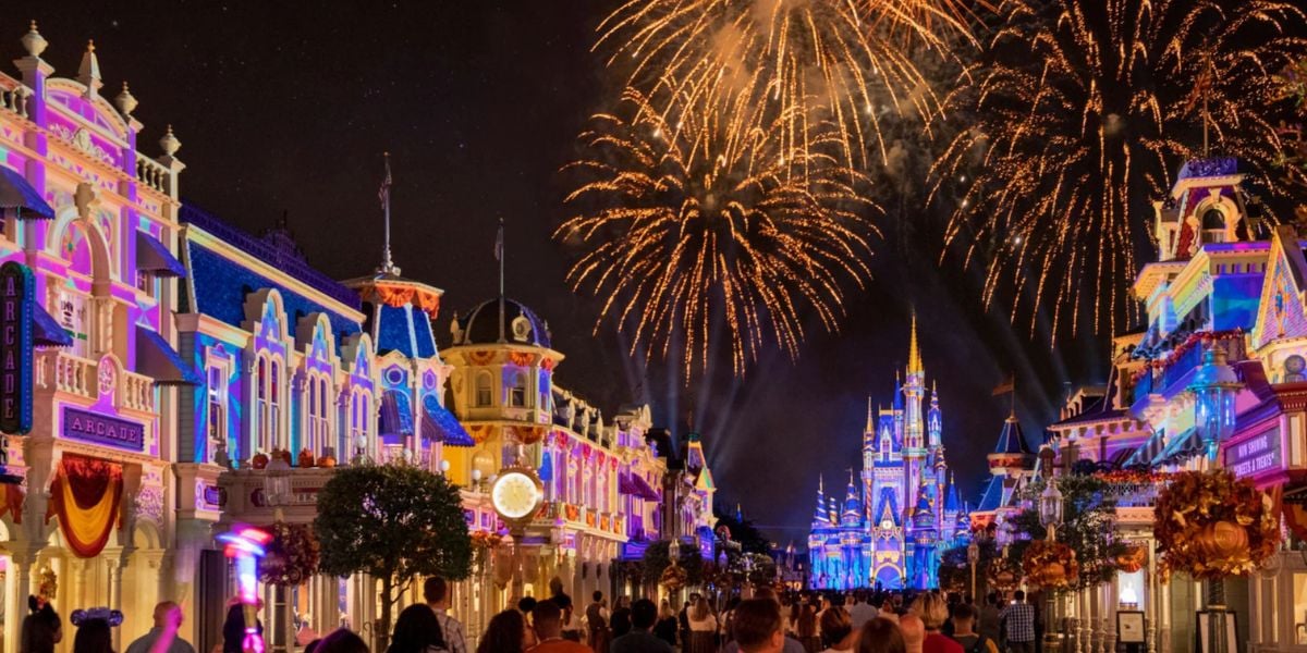Colorful Disney Enchantment fireworks light up the night sky above a brightly illuminated castle and vibrant buildings, while crowds of people walk along a bustling street filled with lights and decorations.