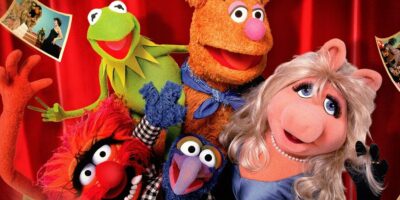 The Muppets characters Animal, Kermit the Frog, Fozzie Bear, Gonzo, and Miss Piggy pose together, smiling in front of a red curtain with photos pinned to it, capturing the spirit of The Muppet Show.