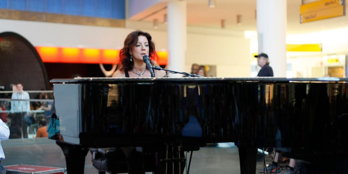 A woman with dark hair sits and sings at a grand piano in a brightly lit indoor public space, with people and signs visible in the background.