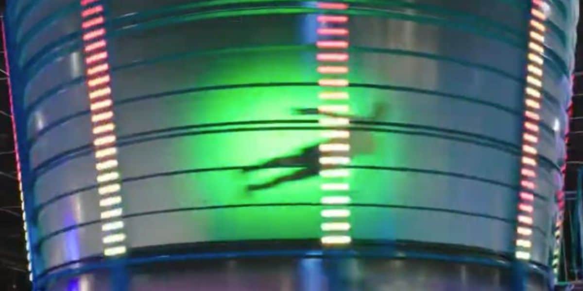 A blurred silhouette of a person is spinning inside a brightly lit, cylindrical amusement ride with horizontal neon lights and a green glow in the background.