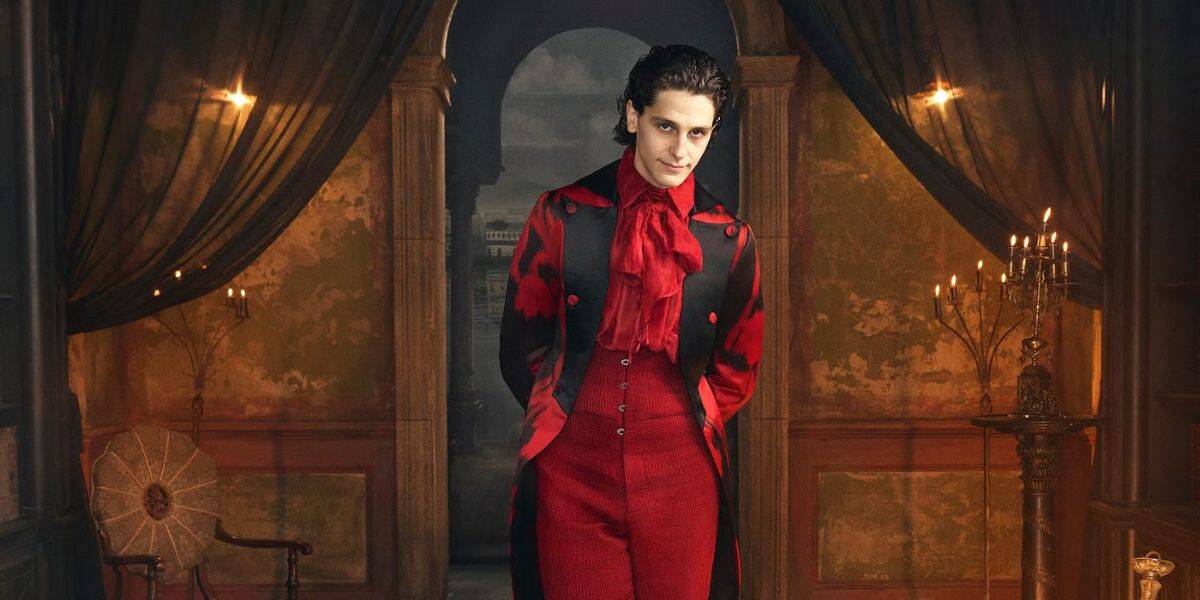 A person with slicked-back dark hair wears a red and black Victorian-style suit with a ruffled red shirt, standing in an ornate, dimly-lit room with tall pillars, draped curtains, and candelabras.