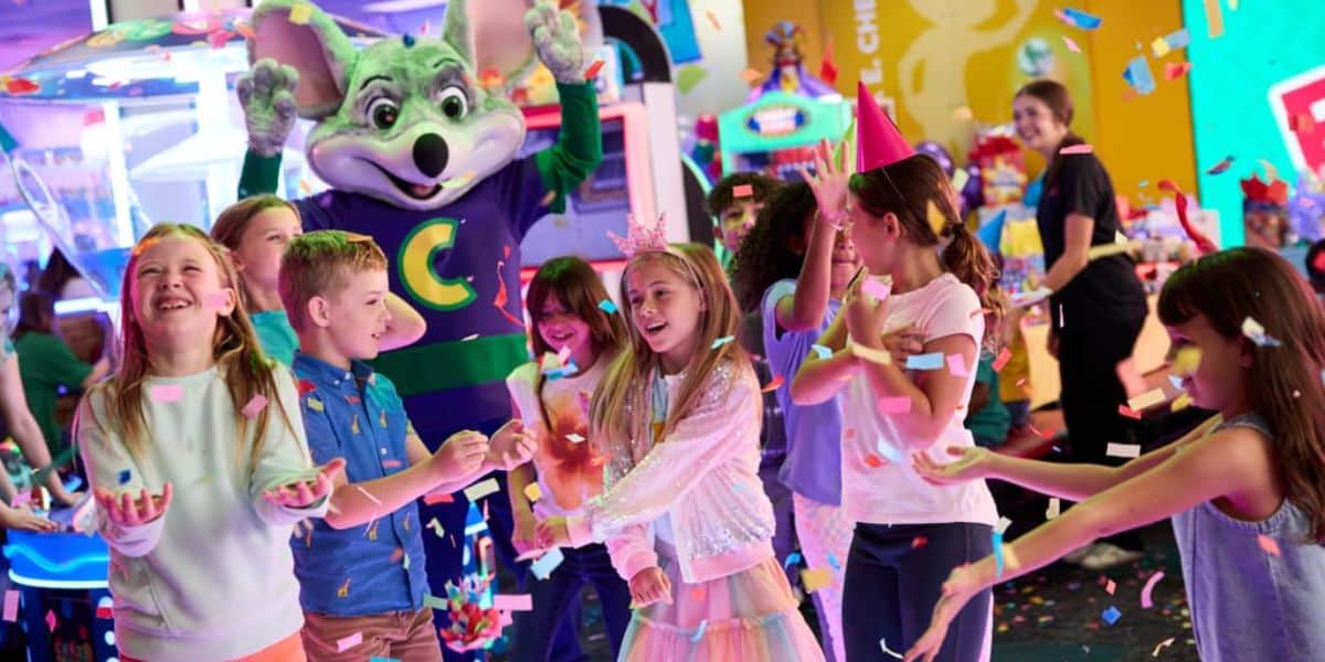 Children celebrate at an arcade with colorful confetti, laughing and dancing as a person in a green mouse mascot costume stands behind them, raising its hands. Bright decorations and arcade games are visible in the background.
