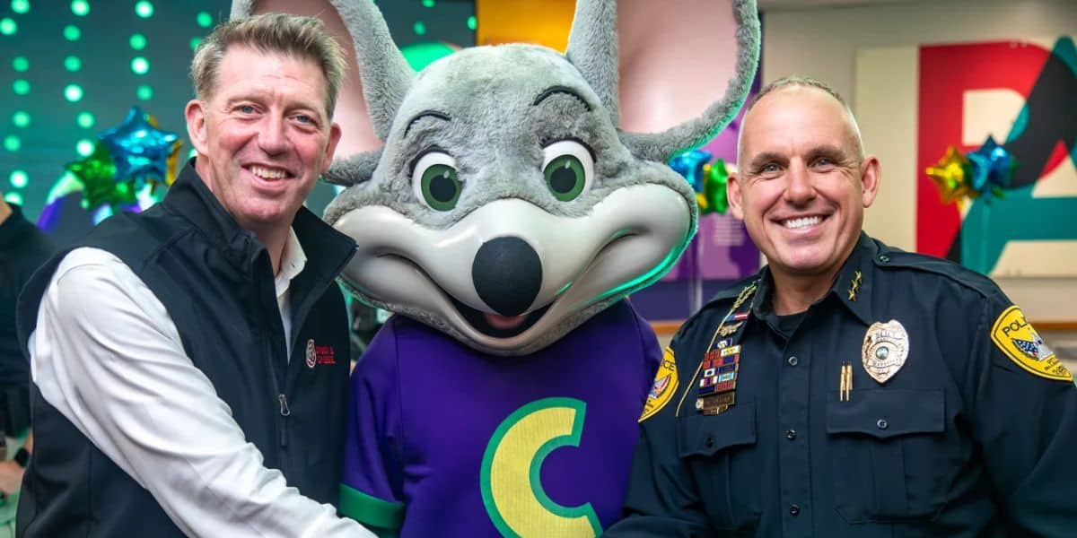 Two men, one in a black vest and one in a police uniform, smile and shake hands while standing beside a person in a gray mouse mascot costume with a big "C" on the shirt. Colorful decorations are in the background.