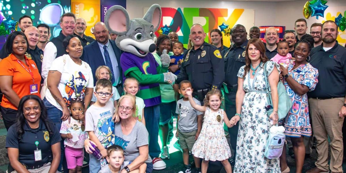 A large group of adults and children, including police officers and a person in a Chuck E. Cheese costume, pose and smile together indoors at a colorful party or community event.