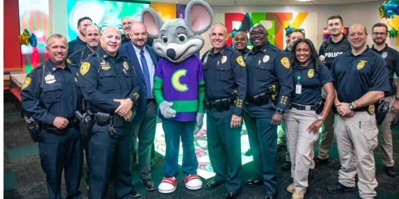 A group of police officers and staff pose together, smiling, around a person in a Chuck E. Cheese mouse costume inside a brightly decorated room.