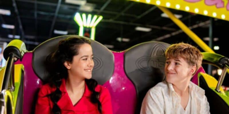 Two teenagers sit side by side on a brightly colored amusement park ride, smiling at each other. Neon lights and a fun, lively atmosphere can be seen in the background.