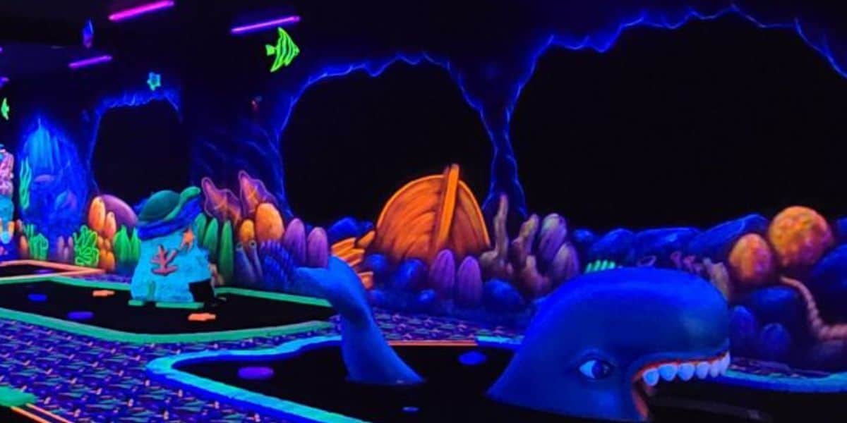 A glow-in-the-dark mini-golf course with an underwater theme, featuring bright neon colors, a whale obstacle, fish, seaweed, and sea caves illuminated by black lights.