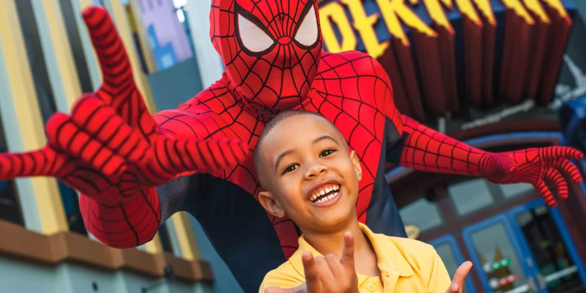 A child smiles and poses with Spider-Man, who is making a web-shooting gesture in front of a colorful theme park backdrop.