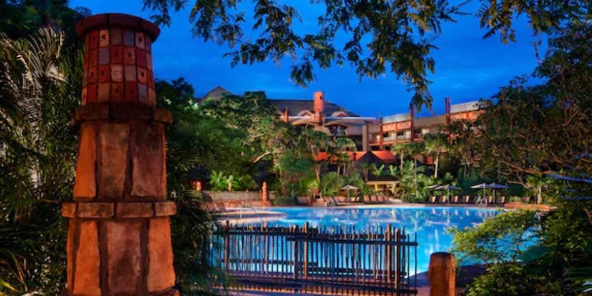 A serene, illuminated swimming pool surrounded by lush greenery and rustic stone pillars evokes the tranquil ambiance of Animal Kingdom Lodge, with a resort building in the background under a twilight sky.