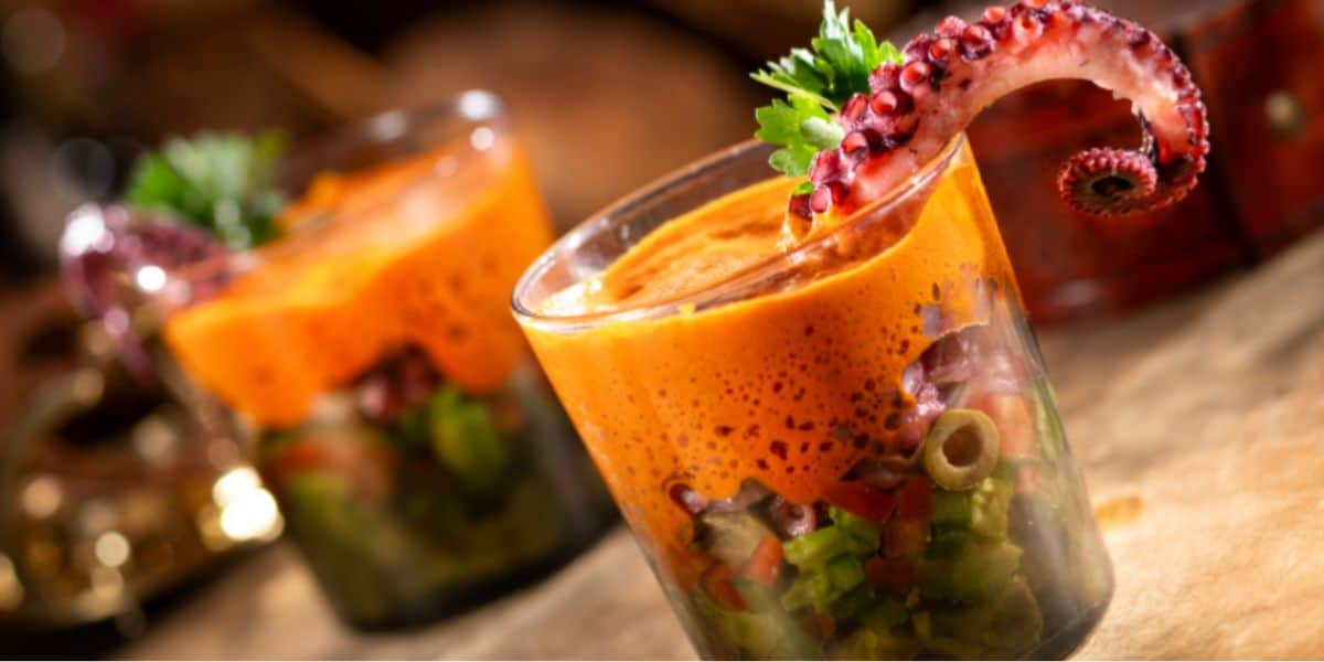 Two glasses filled with a colorful chopped vegetable salad, topped with an orange foam and garnished with an octopus tentacle and fresh herbs—a dish that looks straight out of a Disney-inspired feast.