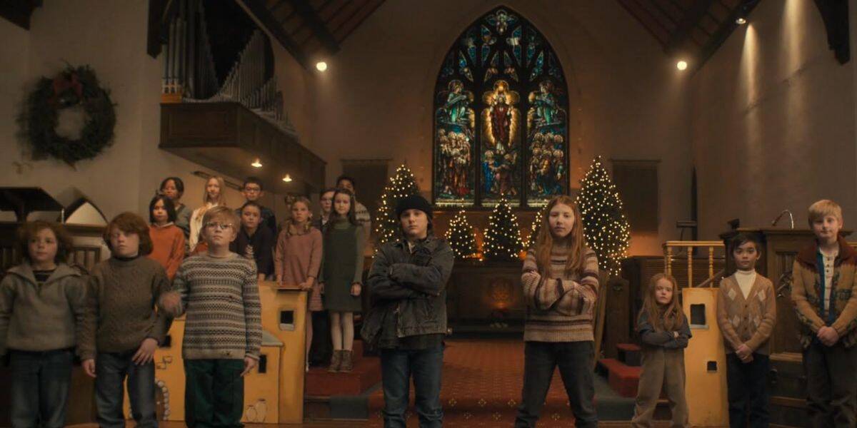 A group of children, dressed in winter clothes, stands in rows inside a church decorated with Christmas trees and a large stained-glass window—capturing a scene reminiscent of tom bancroft’s heartwarming illustrations.