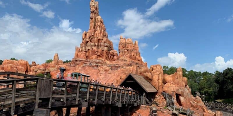 A roller coaster train speeds along wooden tracks on Big Thunder Mountain Railroad in front of red rock formations under a blue sky with scattered clouds, surrounded by trees and greenery at Disney World.