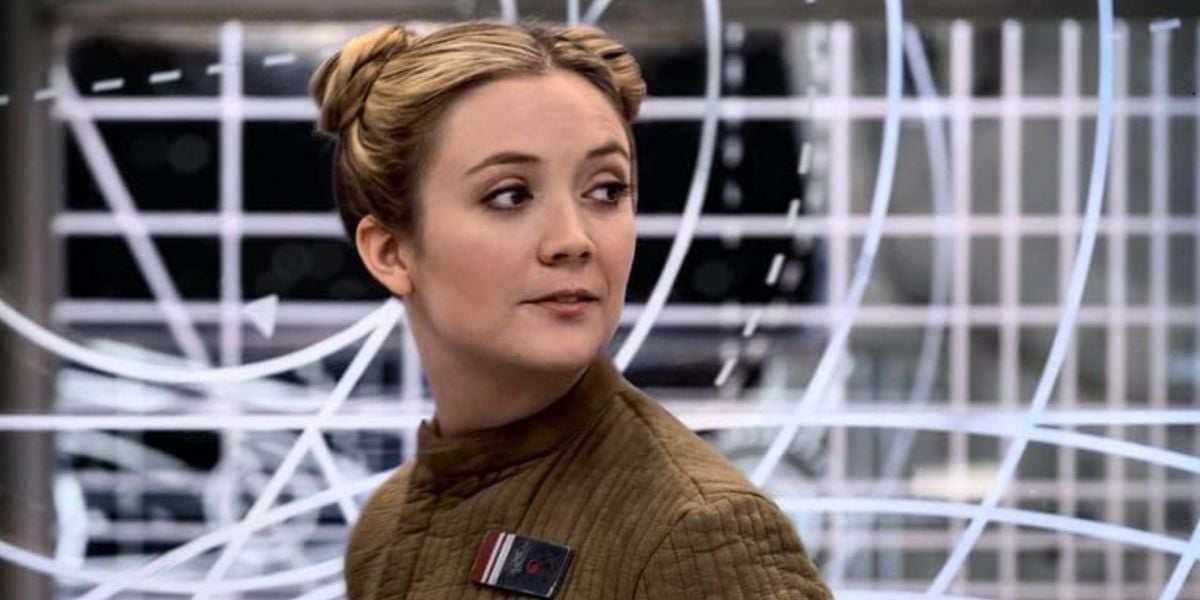 A woman with light brown hair styled in two buns, reminiscent of Carrie Fisher, wears a brown uniform with a badge and stands before a large, futuristic display screen featuring white lines and grid patterns.