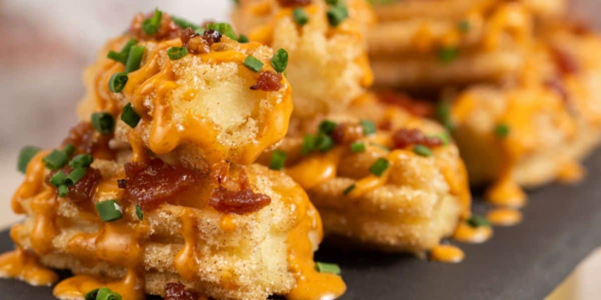 Close-up of crispy, golden potato bites topped with creamy orange cheese sauce, chopped chives, and bits of bacon, served on a dark plate—perfect for bringing a touch of Disney magic to your snack time.