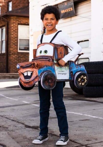 A smiling child stands outdoors wearing a colorful Disney Tow Mater outfit, the rusty tow truck character from the movie Cars. The costume hangs from the child’s shoulders over jeans and a white shirt.