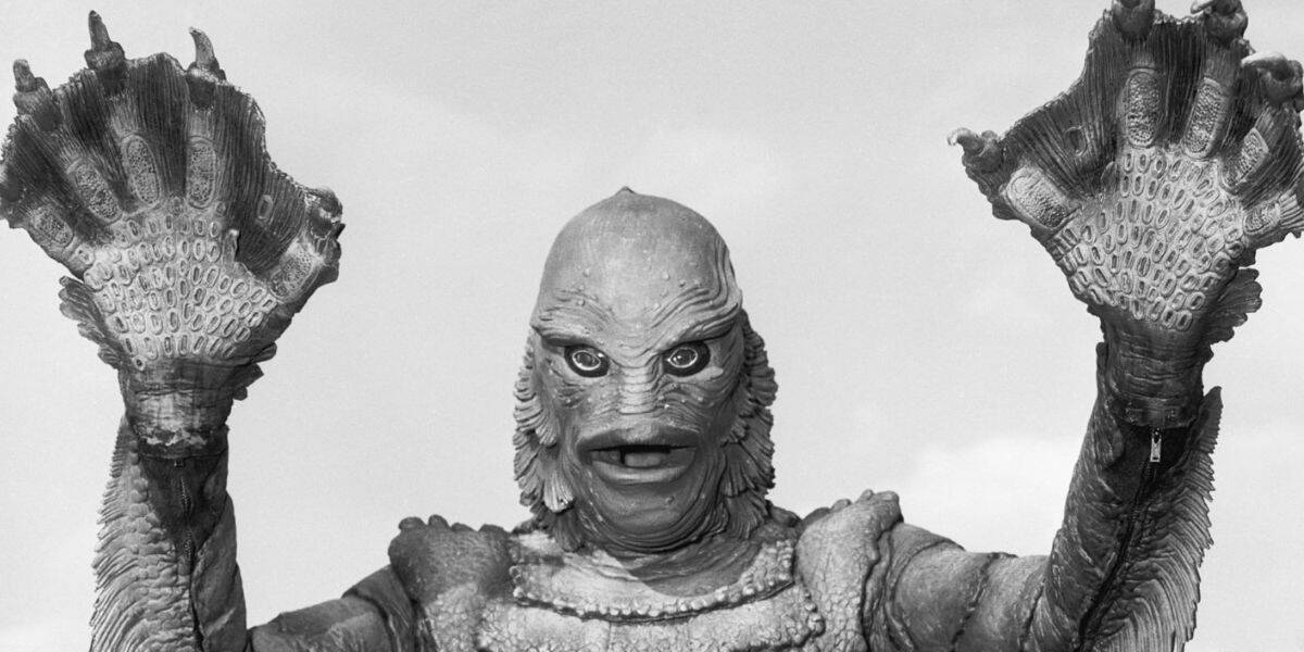 A person in a detailed, universal creature costume with scaly skin, large webbed hands, and bulging eyes raises their arms against a light sky background.