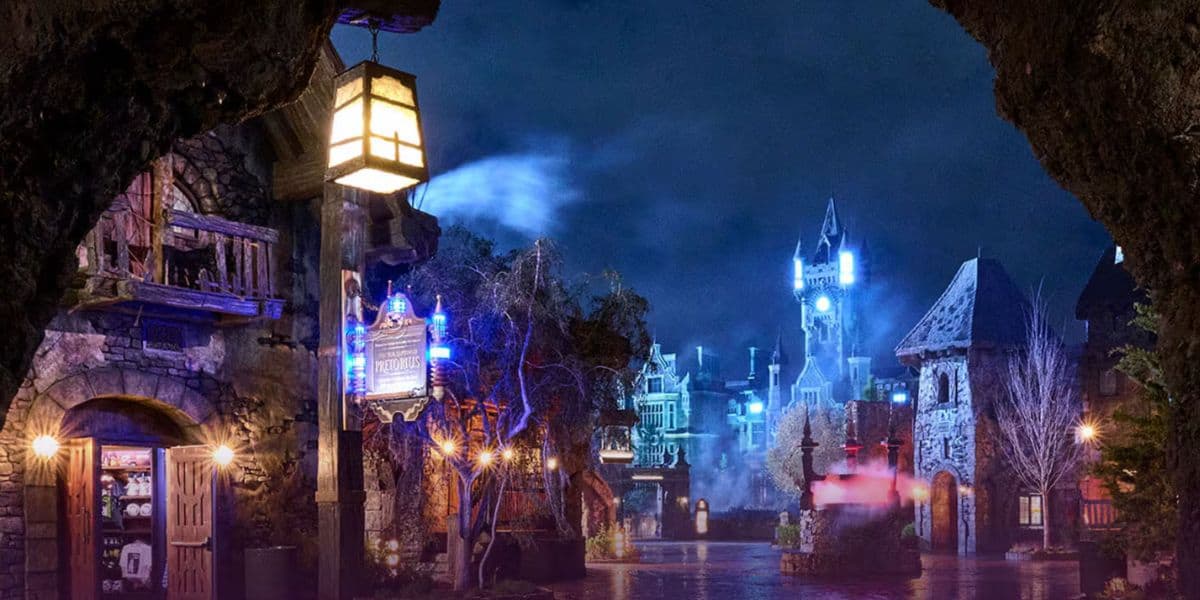 A universal nighttime scene of a fantasy village with stone buildings, glowing blue lights, and a distant castle. Bare trees and drifting mist create a mysterious atmosphere, while a lantern hangs in the foreground.