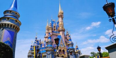 Cinderella Castle at Disney World, decorated in blue and gold with flags and spires, stands against a bright blue sky with scattered clouds—an iconic sight that helps guests forget any Disney problems they may encounter. Surrounding turrets and lampposts frame the scene.