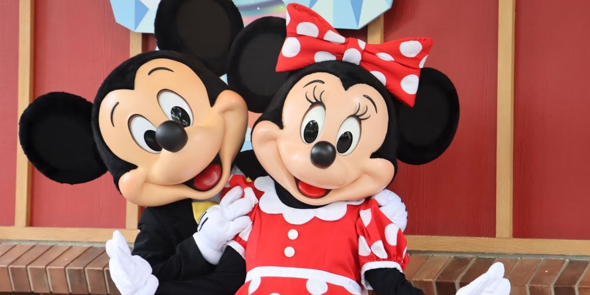 Two people dressed as Mickey Mouse and Minnie Mouse pose together, smiling. With Minnie’s polka dot dress and Mickey’s bow tie, they capture the joy of Disney World Park Expansion in front of a vibrant red wall.