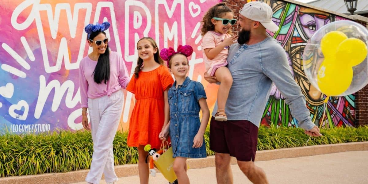 A Disney-loving family of five, smiling and wearing colorful clothing and Mickey Mouse ears, walks in front of a vibrant mural that reads “WARM.” One man holds a child and a Mickey balloon, while two girls and a woman walk beside them.