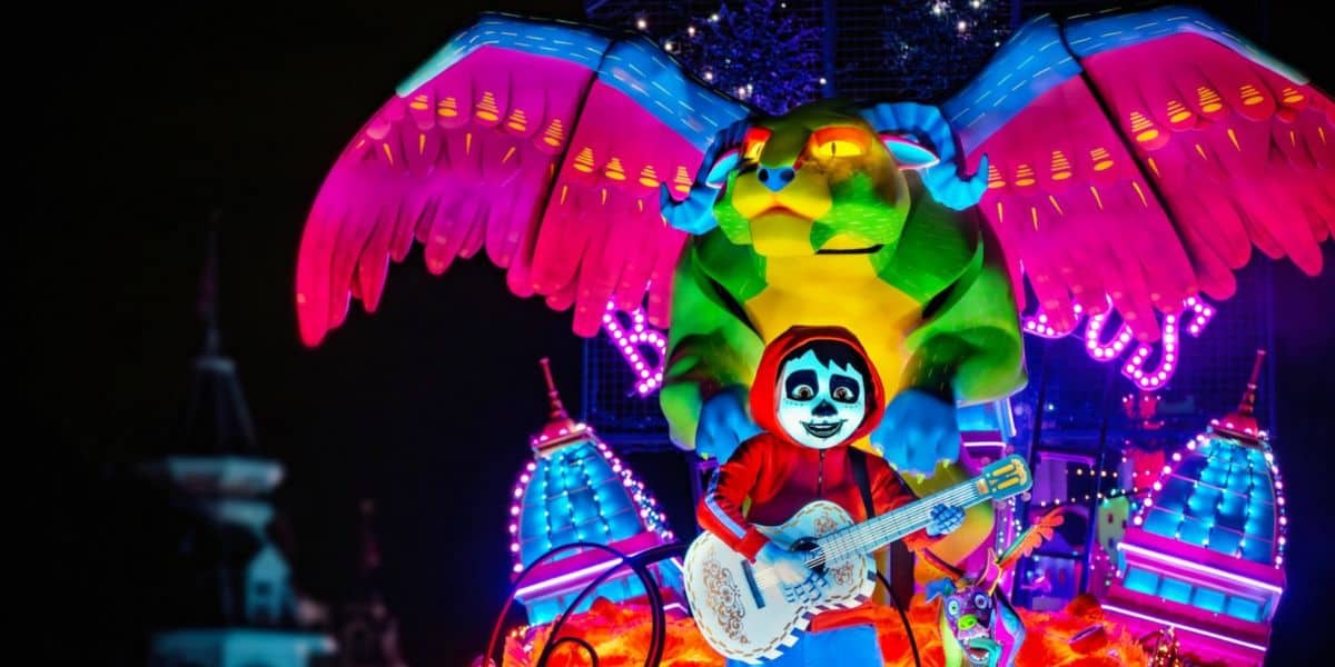 A vibrant parade float features a child in a red hoodie and white face paint playing guitar, with a large, colorful winged creature behind them. Bright neon lights and bold colors illuminate the lively parade night scene.