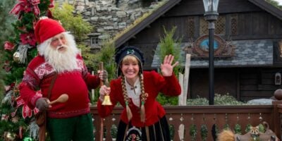 A man and woman in festive traditional Nordic costumes stand by a decorated Christmas tree, smiling. Celebrating Christmas, the woman waves and holds a bell, while the man, dressed like a gnome, holds a wooden spoon. A rustic house and greenery are in the background at Disney World.