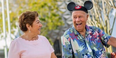An older woman and man smile at each other outdoors. The man sports a colorful Mickey Mouse shirt, matching ears hat, and proudly displays his annual pass, while the woman wears a light pink top. Trees and a dome-shaped structure are in the background.