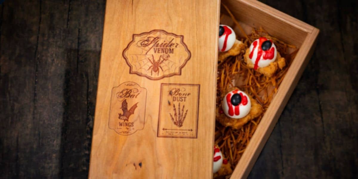 A wooden box with Halloween-themed Disney labels holds spooky treats resembling eyeballs with red syrup and black decorations, nestled on straw-like material atop a dark wooden surface.