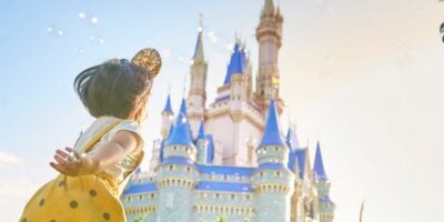 A child in a yellow dress and mouse ears stands with arms outstretched, facing a fairytale castle with blue spires under a bright sky, evoking a sense of wonder and excitement.