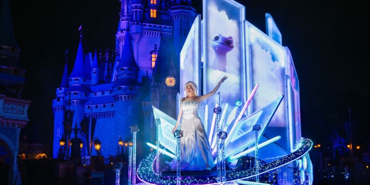 A performer dressed as Elsa from Frozen stands on an illuminated float with icy blue decorations, captivating Disney fans in front of a brightly lit castle at night during one of the magical Disney parades.