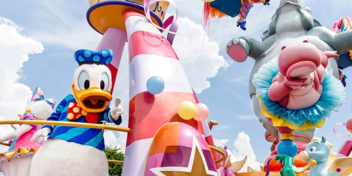 A vibrant parade float at a theme park features Donald Duck in a blue sailor outfit, a pink elephant, and balloons, with other cartoon characters parading in the background beneath a bright blue sky.