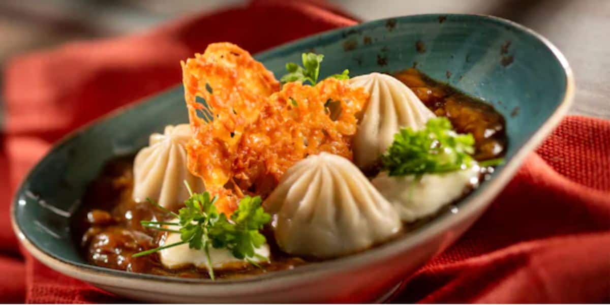 Three steamed dumplings sit in a bowl of savory broth, garnished with fresh parsley and a crispy cheese wafer—an elegant presentation in a teal oval dish on a red cloth napkin, perfect for any food and wine festival experience.