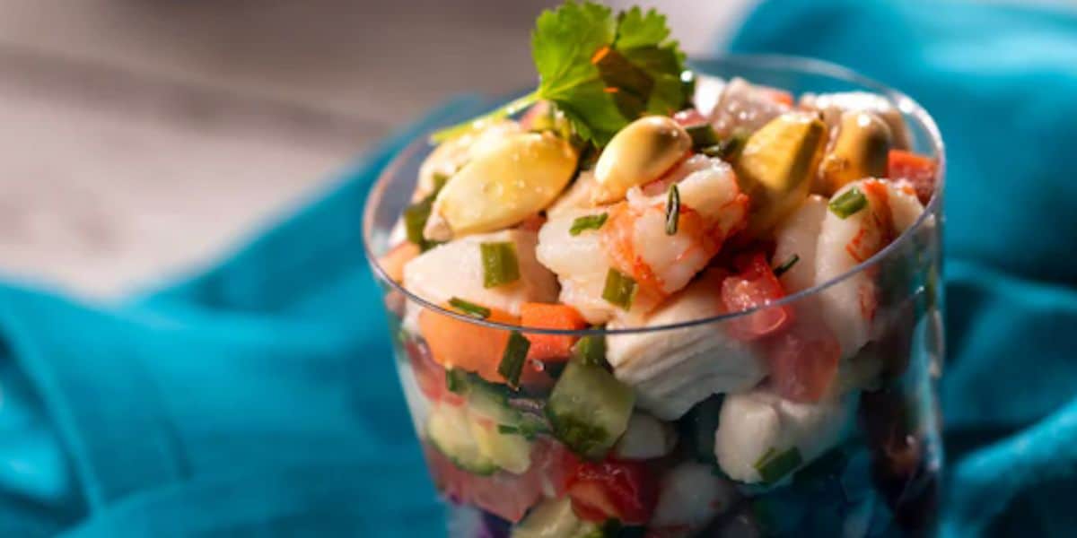 A glass filled with colorful ceviche—fish, shrimp, avocado, tomato, and cucumber—garnished with cilantro and set on a bright blue cloth invites you to savor the flavors of a lively food and wine festival.