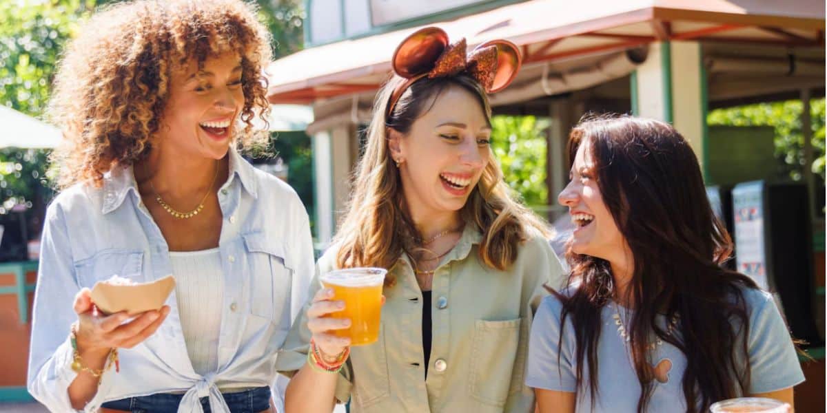 Three women stand outside, laughing and smiling together at a food and wine festival. One holds food, another has a drink and wears Mickey Mouse ears, while the third joins in the fun. They appear to be enjoying a sun-filled day out.