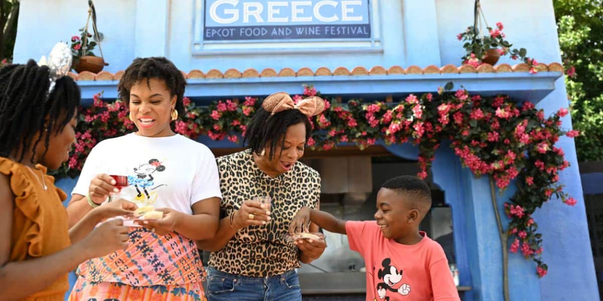 Four people enjoy delicious dishes together in front of a blue booth decorated with flowers and a "Greece Epcot Food and Wine Festival" sign, smiling and soaking up the fun at this lively food and wine festival.