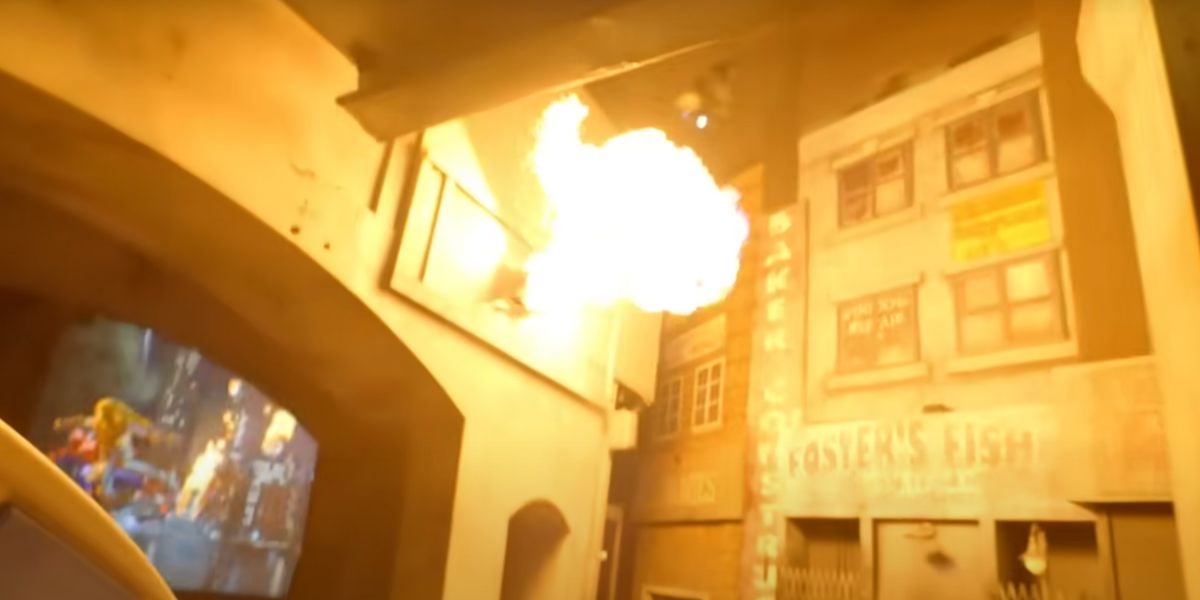 A bright fireball bursts from a building facade in an urban alleyway, with signs and graffiti on the walls and a Spider-Man video projection lighting up the background.