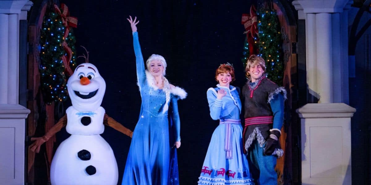 Three people in "Frozen" costumes—Olaf the snowman, Elsa in a blue dress, Anna in a blue skirt, and Kristoff in a winter outfit—pose and smile onstage between decorated christmas trees.