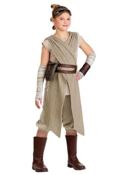 A child in a Rey costume from Star Wars, part of the Disney Costumes collection, wears tan and beige robes, a brown belt and boots, arm wraps, and goggles on their head, standing and smiling against a white background.