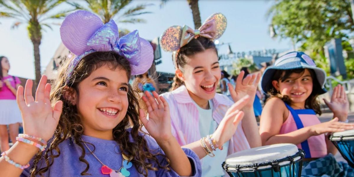 Three smiling children wearing colorful Disney mouse ear headbands and bracelets play drums and raise their hands outdoors on a sunny day, with palm trees and other people in the background.