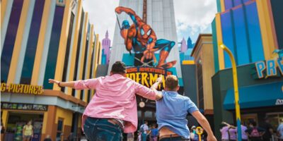 Two people run excitedly toward the entrance of a Spider-Man attraction at a colorful theme park, eager to experience the thrilling world of Spider-Man beneath a giant graphic of the iconic superhero.