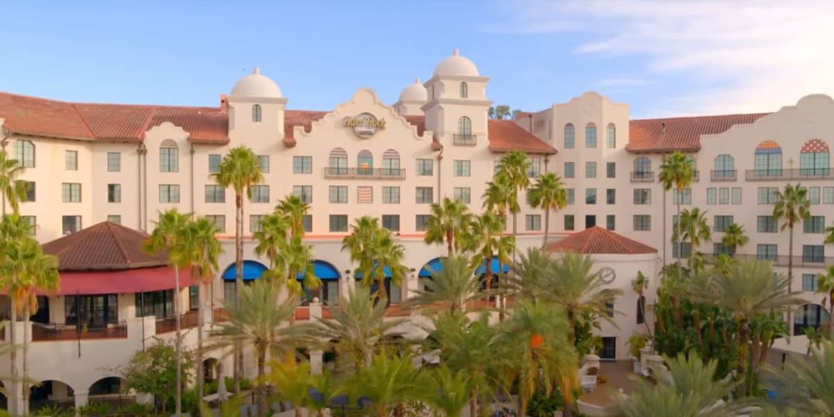 A large white resort hotel with red tile roofs, arched windows, and decorative domes is surrounded by tall palm trees under a blue sky with scattered clouds.